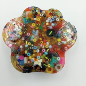 May include: A paw-shaped resin paperweight filled with colourful beads, glitter, and small decorative elements. The resin is clear, allowing the vibrant contents to be visible. The paw print design is a unique and eye-catching decorative item.