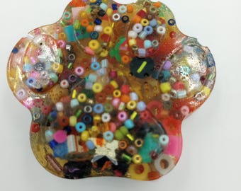 May include: A paw-shaped resin paperweight filled with colorful beads, glitter, and small decorative elements. The resin is clear, allowing the vibrant contents to be visible. The paw print design is a unique and eye-catching decorative item.