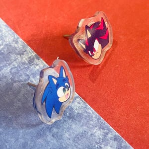 May include: Two clear, irregular-shaped stud earrings. One features a blue cartoon character with a smiling face, and the other has a black and red cartoon character. The earrings are set against a red and blue background.