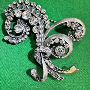May include: Silver-toned brooch with a floral and scroll design, embellished with clear, sparkling rhinestones. The brooch features a pin closure on the back and is set against a green background. A vintage-inspired jewelry piece.