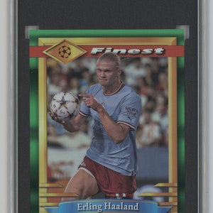 May include: A green and gold framed trading card featuring a photo of Erling Haaland, a soccer player, holding a soccer ball. The card is from the 2022-23 Topps Finest Flashbacks UCC series and is numbered #100. The card is graded 9 by SGC.