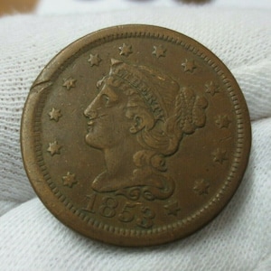 May include: A copper-colored 1853 Indian Head one-cent coin with a portrait of Liberty on the obverse. The coin has a ring of 13 stars around the edge.