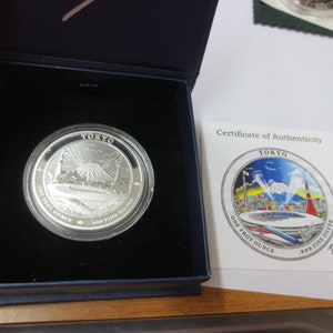 May include: A silver coin in a blue box, featuring the word "TOKYO" at the top. The coin has an image of a mountain and a bridge. A certificate of authenticity is next to the coin, with the words "TOKYO" and "ONE TROY OUNCE".