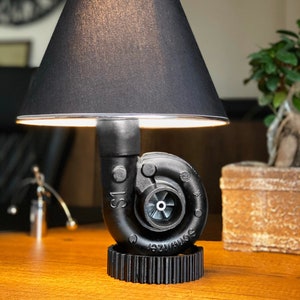 May include: A black table lamp with a unique design, featuring a turbocharger base and a black lampshade. The lamp sits on a gear-shaped base and is placed on a wooden surface. The lamp's height is approximately 12 inches.