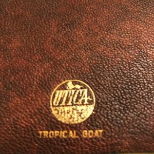 May include: A brown leather journal with a gold embossed circle logo that says "UTICA SHIRTS" and the text "TROPICAL GOAT" below the logo.