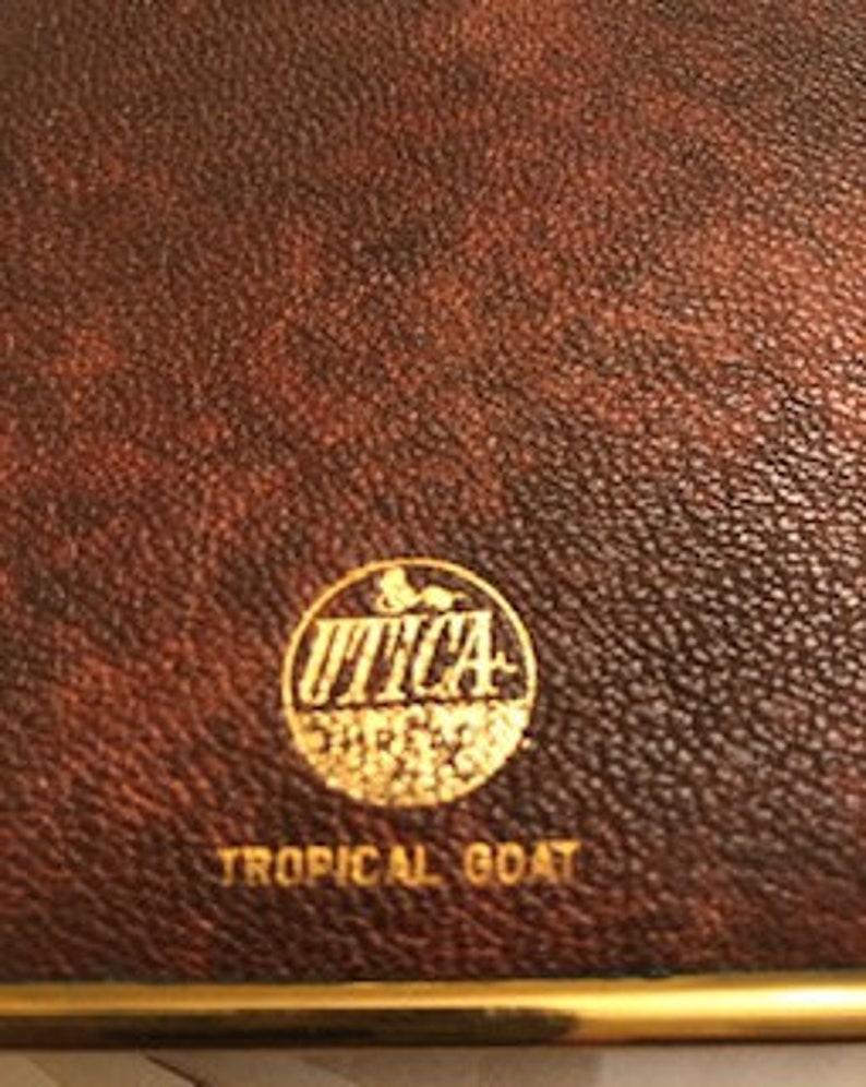 May include: A brown leather journal with a gold embossed circle logo that says "UTICA SHIRTS" and the text "TROPICAL GOAT" below the logo.