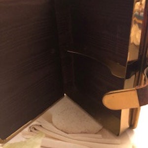 May include: A small, rectangular, gold-colored box with a brown leather exterior. The box is open, revealing a soft, white interior. The box has a gold-colored latch and a brown leather strap.