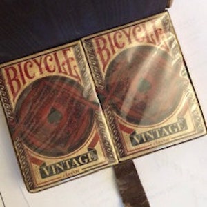 May include: Two decks of Bicycle Vintage playing cards in their original box. The box is brown leather with a gold clasp. The cards feature a vintage design with a red, white, and blue color scheme.