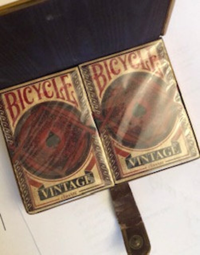 May include: Two decks of Bicycle Vintage playing cards in their original box. The box is brown leather with a gold clasp. The cards feature a vintage design with a red, white, and blue color scheme.