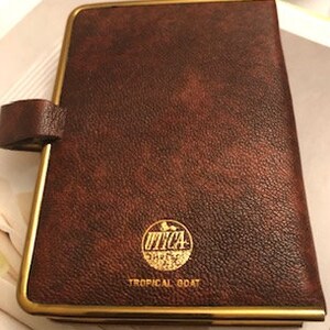 May include: A brown leather-bound journal with a gold trim and a gold embossed logo that says "UTICA" with a picture of a tropical bird. The text "TROPICAL BOAT" is printed below the logo.