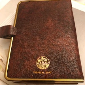 May include: A brown leather-bound journal with a gold trim and a gold embossed logo that says "UTICA" with a picture of a tropical bird. The text "TROPICAL BOAT" is printed below the logo.