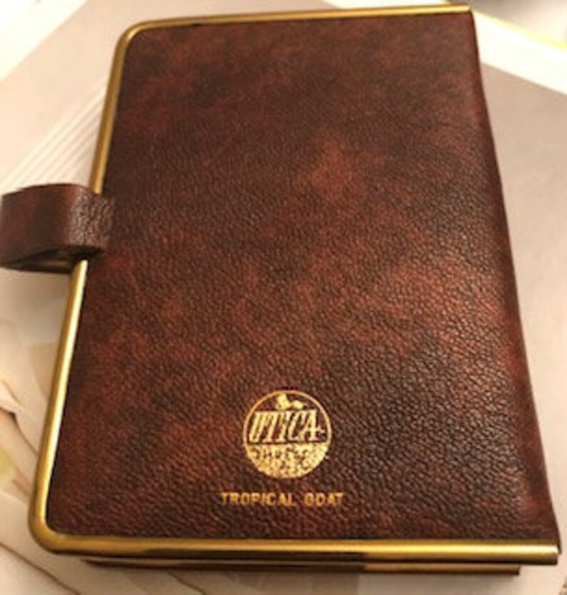 May include: A brown leather-bound journal with a gold trim and a gold embossed logo that says "UTICA" with a picture of a tropical bird. The text "TROPICAL BOAT" is printed below the logo.