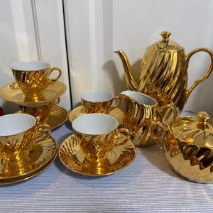 May include: A complete gold tea set, including a teapot, creamer, sugar bowl, and several teacups and saucers. The set has a textured, ridged design. The teacups have white interiors. The set is arranged on a light blue and white striped surface.