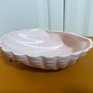 May include: A light pink ceramic seashell-shaped bowl. The bowl has a scalloped edge and a smooth, glossy interior. The bowl is sitting on a brown surface.