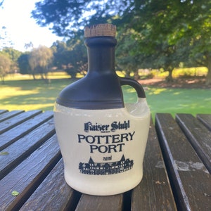 May include: A white ceramic bottle with a black cork stopper and a black handle. The bottle is labelled "Kaiser Stuhl Pottery Port 750 ml".