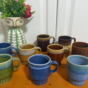 May include: A collection of nine ceramic mugs in various colours including blue, green, brown, and yellow, featuring ribbed bases and smooth rims. The mugs are arranged in a group, with some displaying two-tone glaze effects and others solid colours.