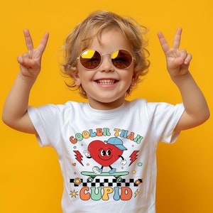 May include: A white t-shirt featuring a colourful graphic of a heart on a skateboard. The heart is wearing a blue hat and has a smiling face. The text "Cooler Than Cupid" is printed on the shirt.