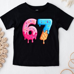 May include: Black short-sleeved t-shirt featuring a colorful graphic of the number "67". The "6" is designed like a pink donut with sprinkles, and the "7" is designed like a blue ice cream cone with a waffle cone base.
