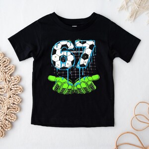 May include: Black short-sleeved t-shirt featuring a graphic design with the number "67" in a soccer ball pattern, dripping with blue paint. The design includes green hands at the bottom. The shirt is likely for a child.