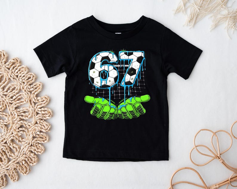 May include: Black short-sleeved t-shirt featuring a graphic design with the number "67" in a soccer ball pattern, dripping with blue paint. The design includes green hands at the bottom. The shirt is likely for a child.