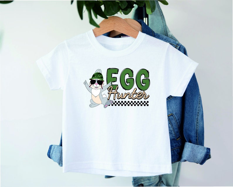 May include: White t-shirt with a cartoon bunny wearing sunglasses and a camouflage hat. The bunny is jumping and the text "EGG Hunter" is printed on the shirt.