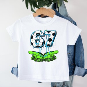 May include: White t-shirt featuring a soccer ball design with the number 67 in black and white, with blue dripping accents. The number is held by green cartoon hands. A fun graphic tee for kids.