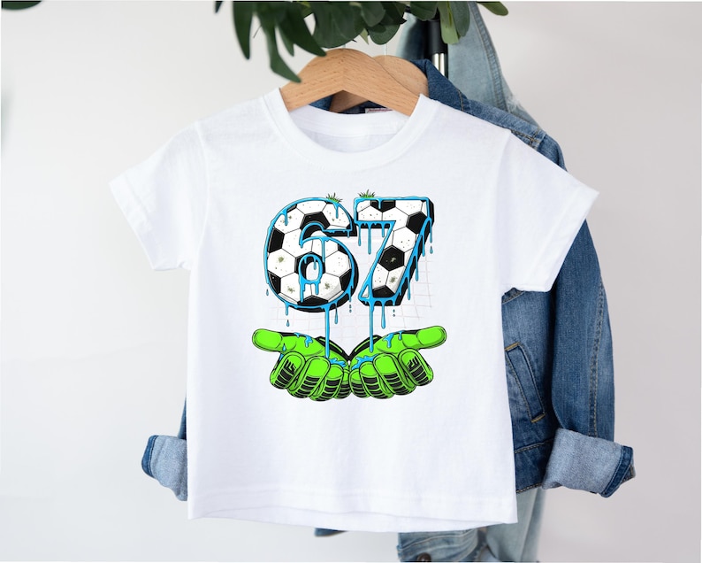 May include: White t-shirt featuring a soccer ball design with the number 67 in black and white, with blue dripping accents. The number is held by green cartoon hands. A fun graphic tee for kids.