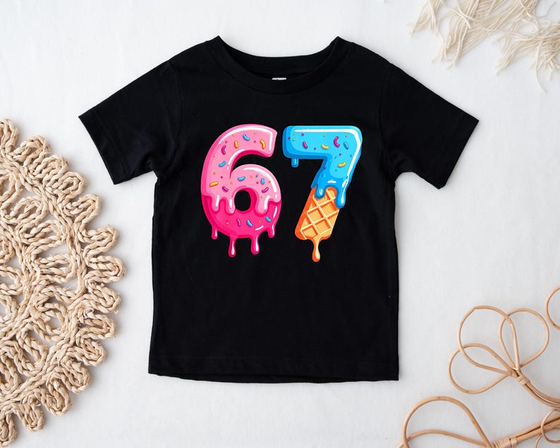 May include: Black short-sleeved t-shirt featuring a colorful graphic of the number "67". The "6" is designed like a pink donut with sprinkles, and the "7" is designed like a blue ice cream cone with a waffle cone base.