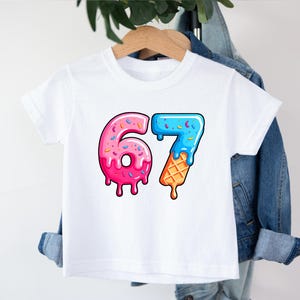May include: White t-shirt with a colorful graphic of the numbers "67". The "6" is designed like a pink donut with sprinkles, and the "7" is blue with sprinkles and an ice cream cone base. The shirt is hanging on a wooden hanger.