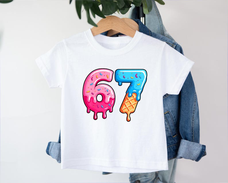 May include: White t-shirt with a colorful graphic of the numbers "67". The "6" is designed like a pink donut with sprinkles, and the "7" is blue with sprinkles and an ice cream cone base. The shirt is hanging on a wooden hanger.