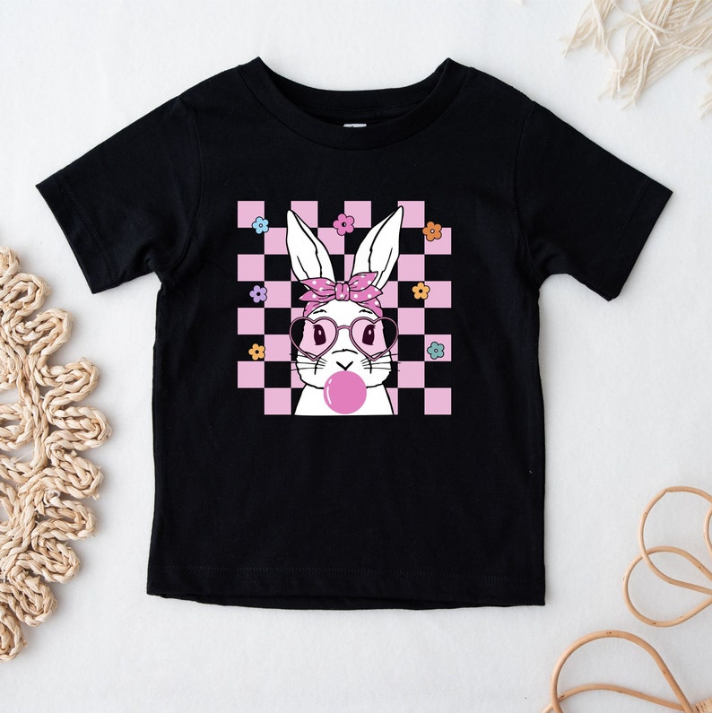 May include: Black t-shirt with a pink and white checkered background featuring a white bunny wearing pink sunglasses and a pink headband, blowing a pink bubble.
