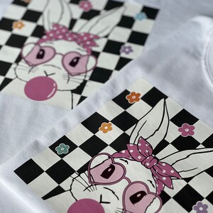 May include: A white bunny with pink heart-shaped sunglasses and a pink polka dot headband is blowing a pink bubble. The bunny is on a black and white chequered background with colourful flowers.