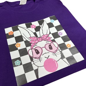 May include: Purple t-shirt featuring a white bunny wearing pink heart-shaped sunglasses and a pink polka dot headband. The bunny is blowing a pink bubble and is set against a black and white checkered background with colourful flowers.
