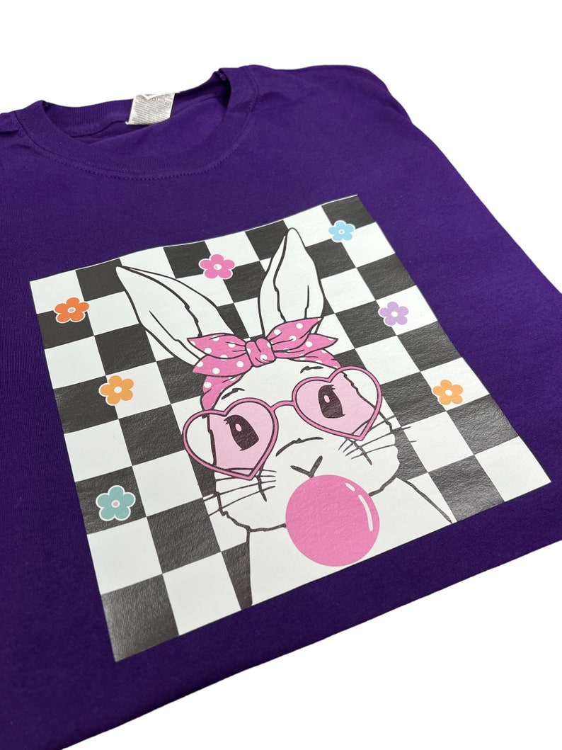 May include: Purple t-shirt featuring a white bunny wearing pink heart-shaped sunglasses and a pink polka dot headband. The bunny is blowing a pink bubble and is set against a black and white checkered background with colourful flowers.
