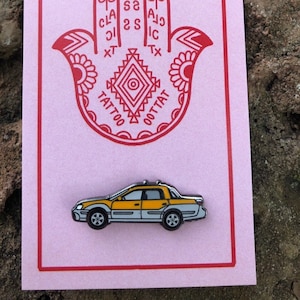May include: A pink card with a red hamsa hand design and the words "Classic Tattoo" in a circle around the hand. A yellow and grey car pin is below the hamsa.