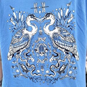 May include: A light blue t-shirt featuring a detailed dark blue graphic print of two large herons facing each other, surrounded by smaller illustrations of a frog, snakes, turtles, dragonflies, cattails, and various small creatures.