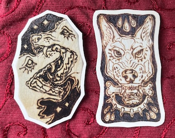 May include: Two wood-burned stickers featuring black and white illustrations of wolf heads. The first sticker shows a wolf head with its mouth open, revealing sharp teeth. The second sticker shows a wolf head with a bone in its mouth and a spiked collar around its neck.