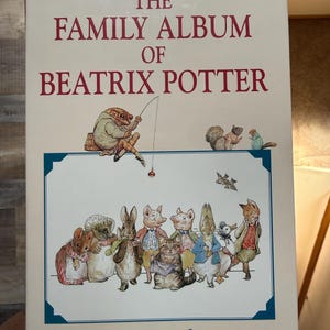 May include: A hardcover book titled The Family Album of Beatrix Potter with text by Abigail Jacobs. The cover features a cream background with red text and illustrations of anthropomorphic animals including a frog fishing and a group of dressed animals.