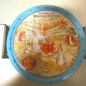 May include: A round metal tin featuring a light blue border with small blue leaf motifs. The center displays an illustration titled THE WORLD OF BEATRIX POTTER, showing various rabbits, squirrels, and a hedgehog in a garden setting with soft colors.