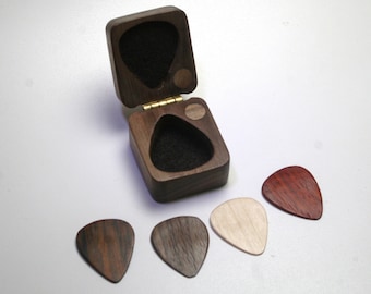 May include: A wooden guitar pick box with a hinged lid, open to reveal a black interior. Four wooden guitar picks in various shades of brown and tan are arranged around the box. The box and picks are on a white surface.