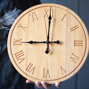 May include: A circular wooden clock featuring Roman numerals. The clock face is light brown, with dark brown Roman numerals and a dark brown border. The clock hands are black. The clock is being held.
