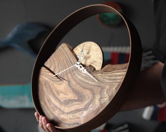 May include: A handcrafted wooden clock with a unique design. The clock features a circular frame and a wooden face with a natural wood grain pattern. The clock hands are silver. The clock is held by a person.
