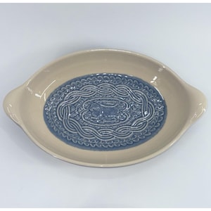 Puede incluir: Una fuente de cerámica ovalada con borde y asas de color crema. El centro presenta un panel azul hundido decorado con un relieve de un conejo rodeado por un borde trenzado y pequeños motivos de hojas en relieve sobre la superficie.