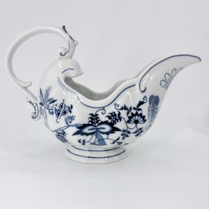 May include: A white porcelain gravy boat featuring a blue onion pattern with intricate floral and botanical motifs. The vessel has a curved handle, a wide pouring spout, and a pedestal base, all decorated with detailed blue painted designs.