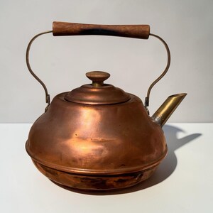 May include: A copper kettle with a wooden handle and lid knob. The spout and handle supports are brass. The kettle has a rounded body and a vintage aesthetic.