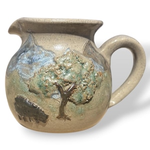 May include: A handmade ceramic pitcher with a textured surface. The pitcher features a raised relief design of a tree, a mountain range, and a dark-colored animal. The colors include beige, green, blue, and brown.