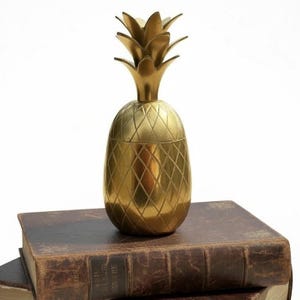 May include: A metallic gold pineapple-shaped container with a textured diamond-patterned surface and a removable lid featuring pointed leaves at the top. The object has a smooth, polished finish and a rounded base, standing upright as a decorative piece.