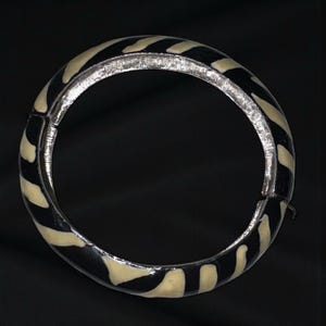May include: A hinged bangle bracelet with a zebra stripe pattern. The bracelet features alternating black and cream-colored stripes. The metal clasp and inner band are silver-toned. The bracelet is shown against a black background.