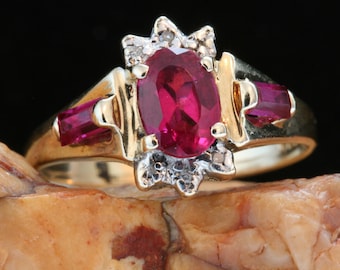 May include: A gold ring featuring a large oval ruby gemstone surrounded by smaller diamonds. The ring has a delicate design with small ruby accents on either side of the main stone.