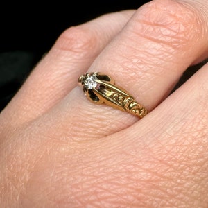 May include: A gold ring with a single diamond set in a decorative setting. The ring is worn on a finger.